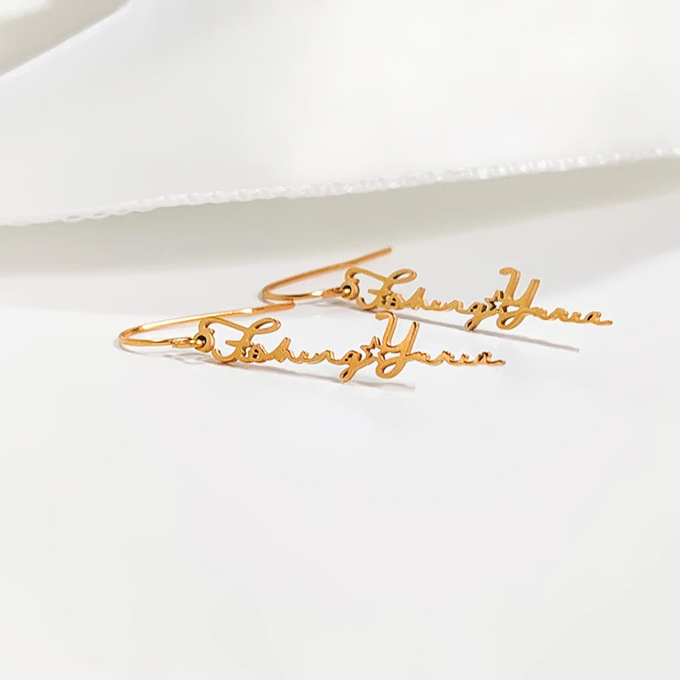 Par de brincos com nome dourado sobre tecido branco