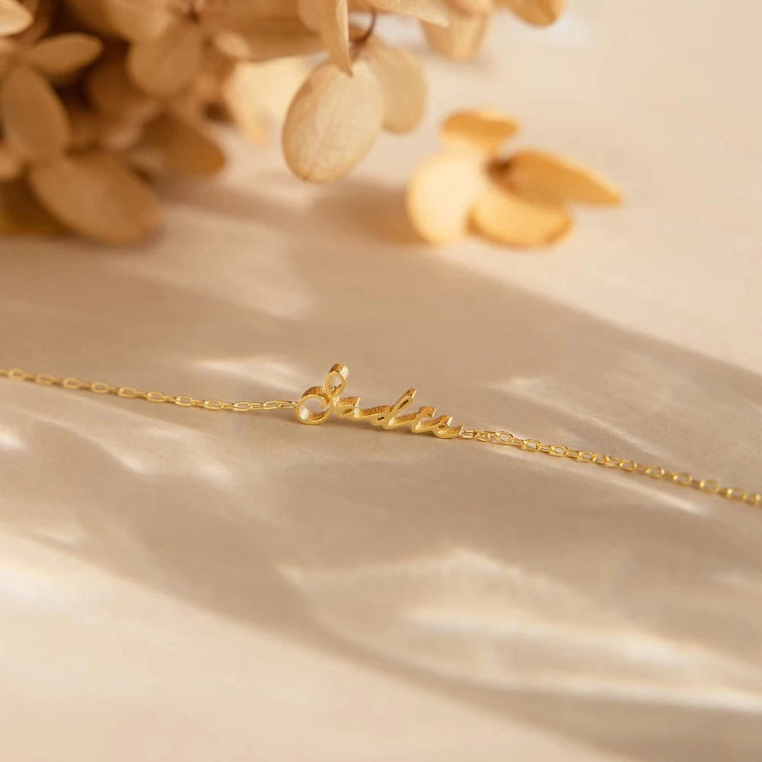 Pulseira com nome dourada em corrente fina sobre tecido claro